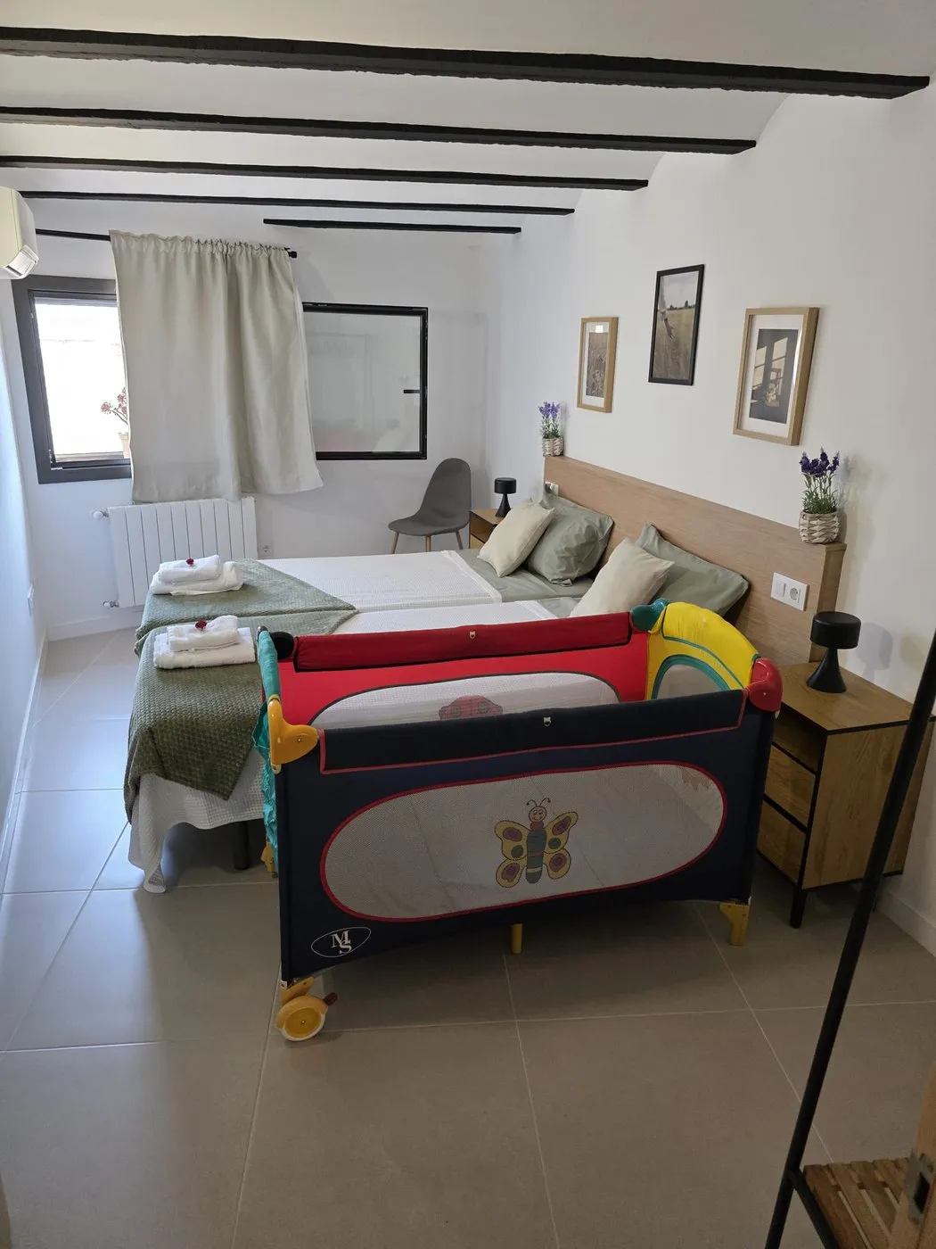 The same twin bedroom with a travel cot set up beside the beds, under the black ceiling beams