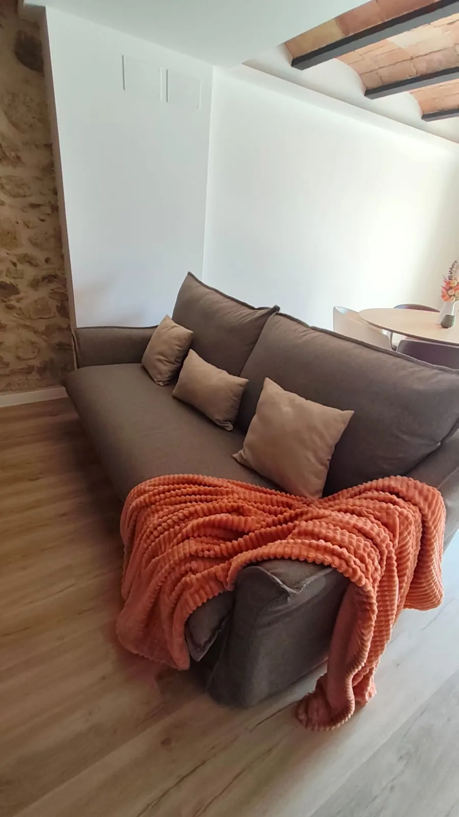 The sofa bed folded away as a sofa, with beige cushions and an orange blanket, next to the stone wall and the round dining table