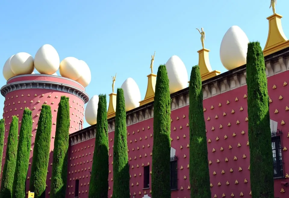 The red façade of the Dalí Theatre-Museum in Figueres, with giant eggs on the roof and a row of cypress trees